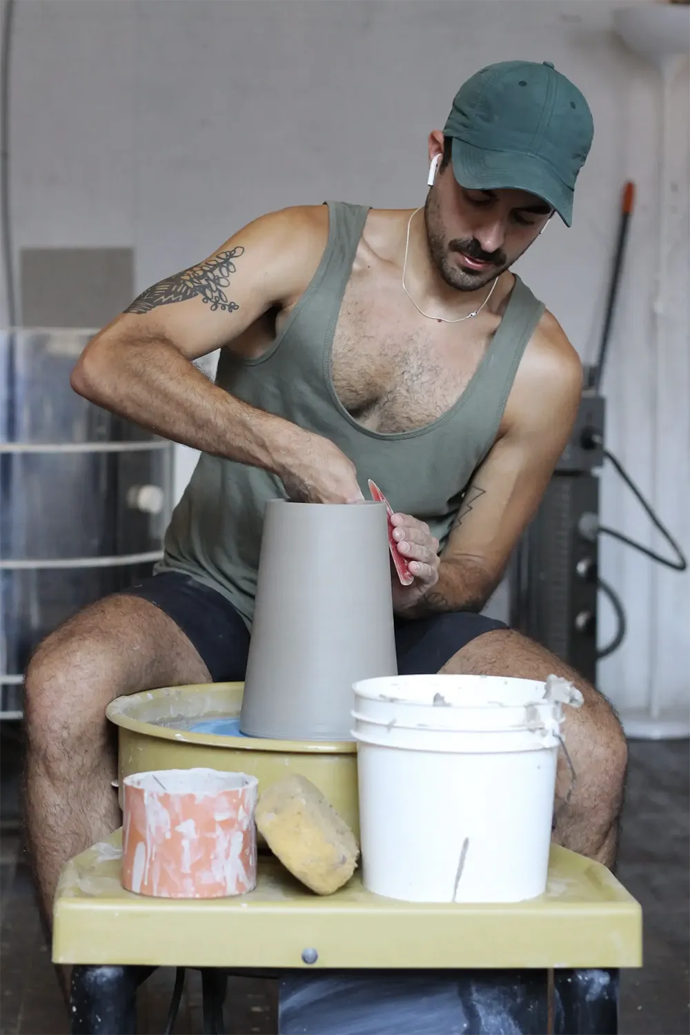 white man wearing a green cap and a sleeveless t-shirt, turning a clay pot