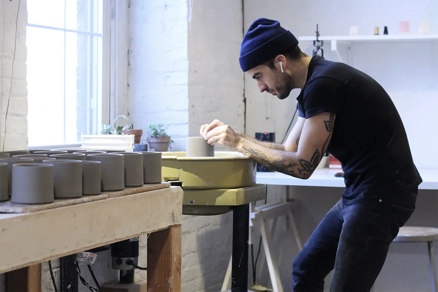 Man wearing black jeans, t-shirt and a blue beanie hat. Sculpting a clay vessel.