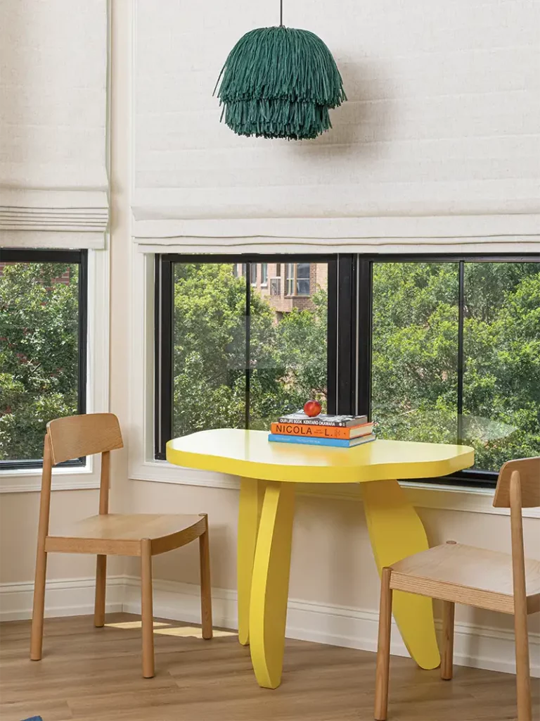 Artistic yellow table next to a large window, with two wooden chairs, books on top, and a hanging green lamp.