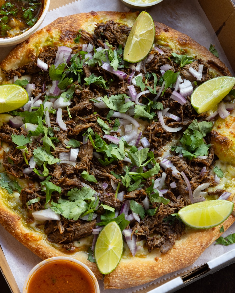 A pizza topped with shredded meat, chopped onions, and fresh cilantro, garnished with lime wedges around the edges, served with small cups of salsa on the side.