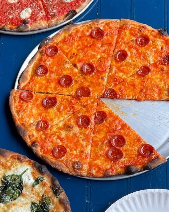 A round pepperoni pizza with one slice removed, showing melted cheese and evenly spaced pepperoni slices, served on a blue table with other dishes nearby.