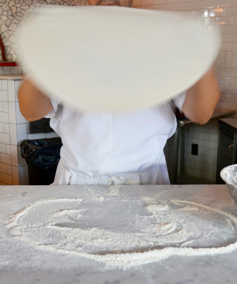 A person stretching a round sheet of pizza dough over a floured countertop in a kitchen, preparing it for toppings.