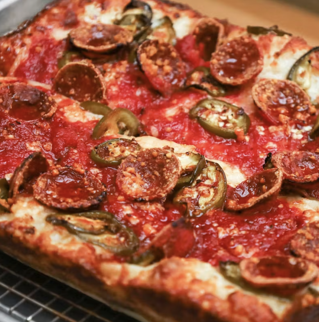 A rectangular pizza topped with melted cheese, slices of pepperoni, halved cherry tomatoes, and sliced jalapeños, resting on a wire rack.