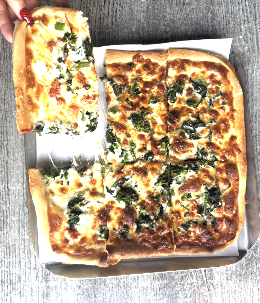 A square pizza in a box topped with melted cheese and scattered greens, with one slice removed and placed to the side, revealing the thick crust and airy interior.