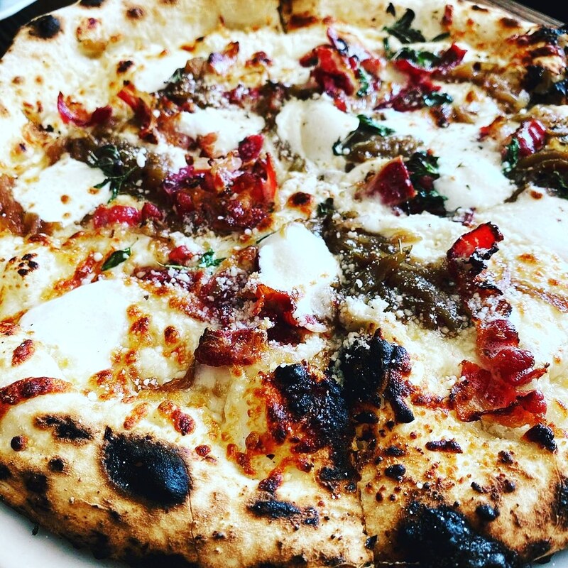 A close-up of a wood-fired pizza with a charred, blistered crust, topped with melted mozzarella, tomato sauce, and pieces of roasted red peppers and herbs.