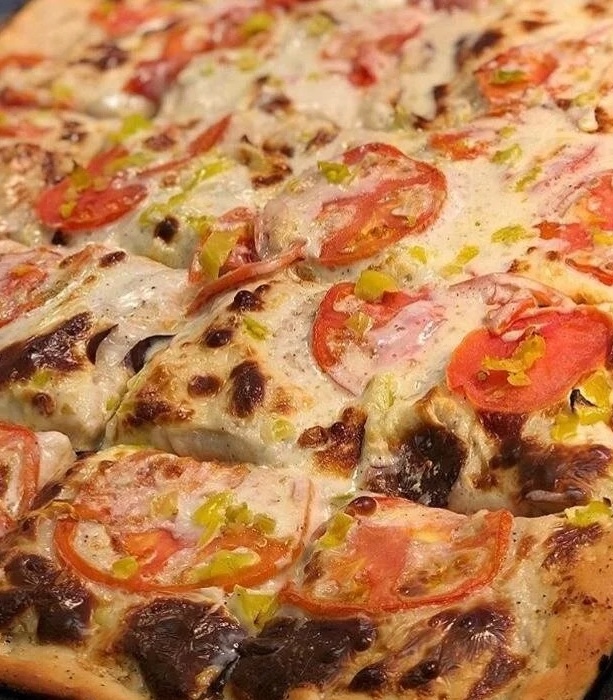A close-up of a pizza with a lightly charred crust, topped with melted cheese, sliced tomatoes, green pepper strips, and pieces of grilled meat.