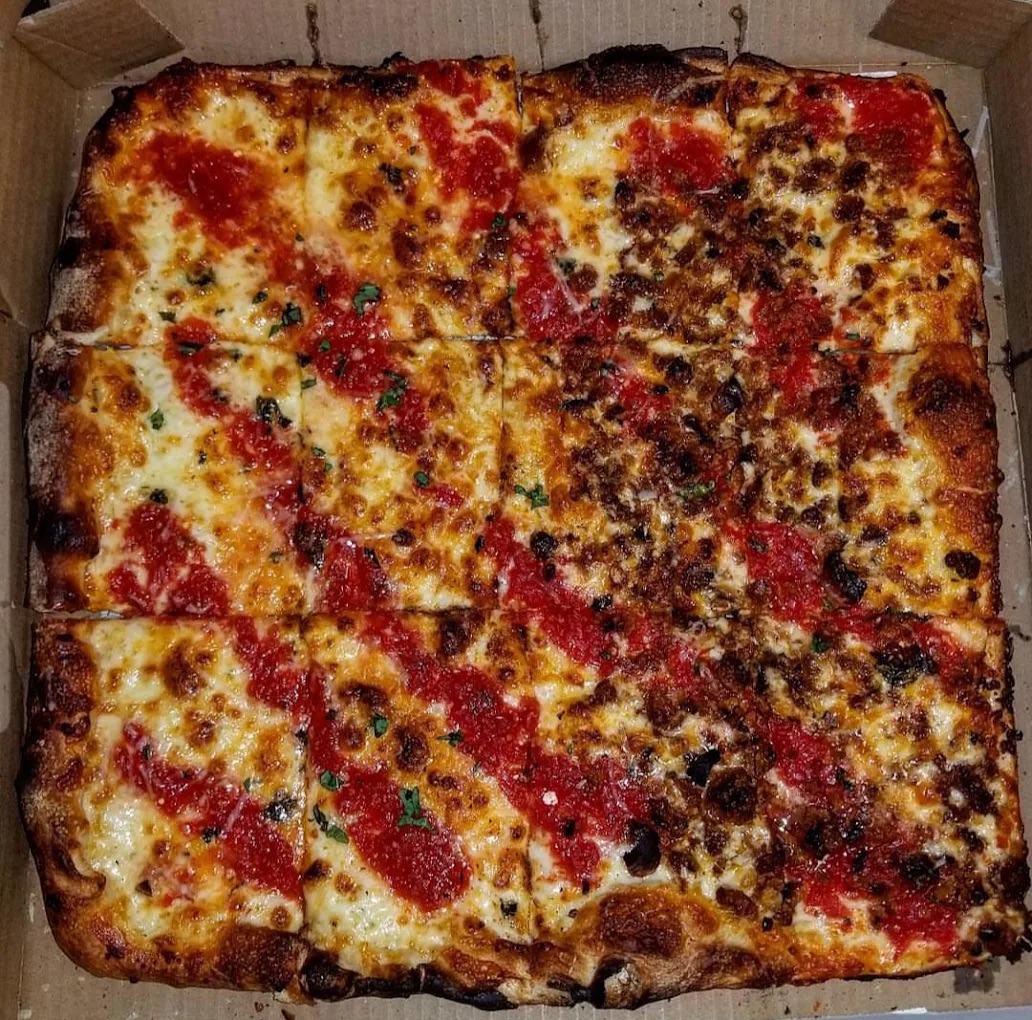 A large rectangular pizza in a box, topped with melted cheese and tomato sauce, with one half covered in crumbled sausage and herbs.