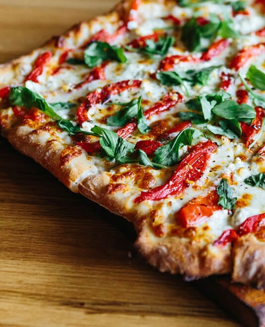 A slice of pizza on a wooden board topped with melted mozzarella, roasted red pepper strips, and fresh basil leaves, with a lightly charred crust.